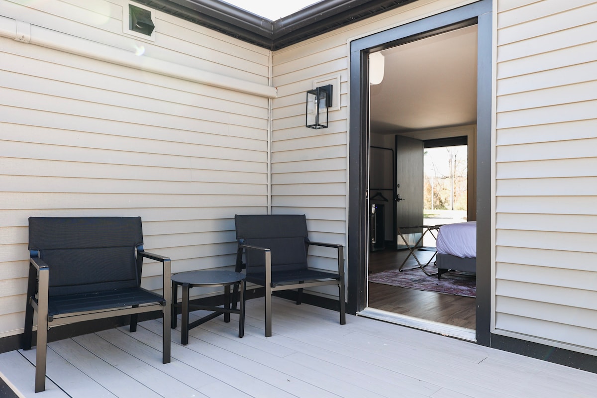 An outdoor seating area features two sleek chairs and a small table, situated on a light-colored deck. The entrance to the indoor space reveals a contemporary interior, with natural light spilling in from the open door.