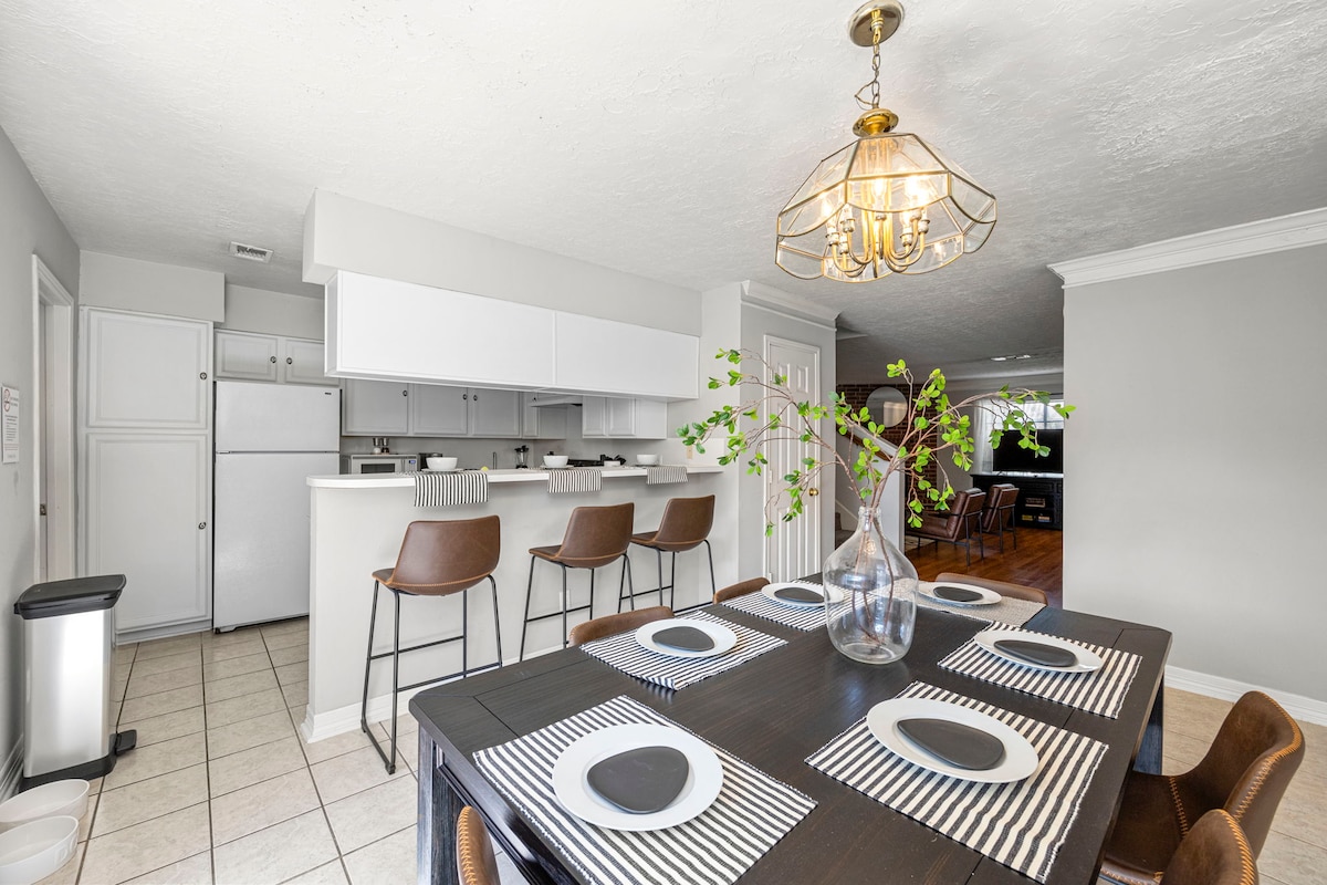 A spacious dining area is highlighted by a dark wooden table set with striped placemats and white plates. Stylish bar stools are positioned along a breakfast bar, with a light fixture featuring a geometric design illuminating the space. A vase with greenery adds a touch of nature.