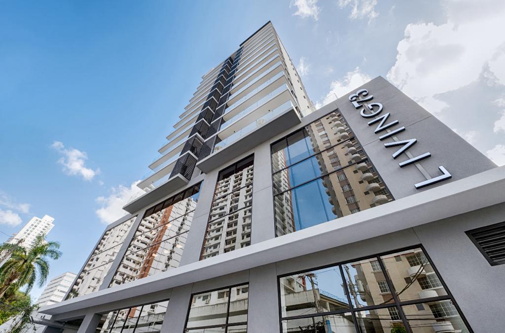 The exterior of the building showcases a modern design with sleek lines and large glass windows. The name 'LIVING' is prominently displayed on the facade. Surrounding palm trees add greenery, and the sky is bright with scattered clouds, creating an open and airy ambiance.