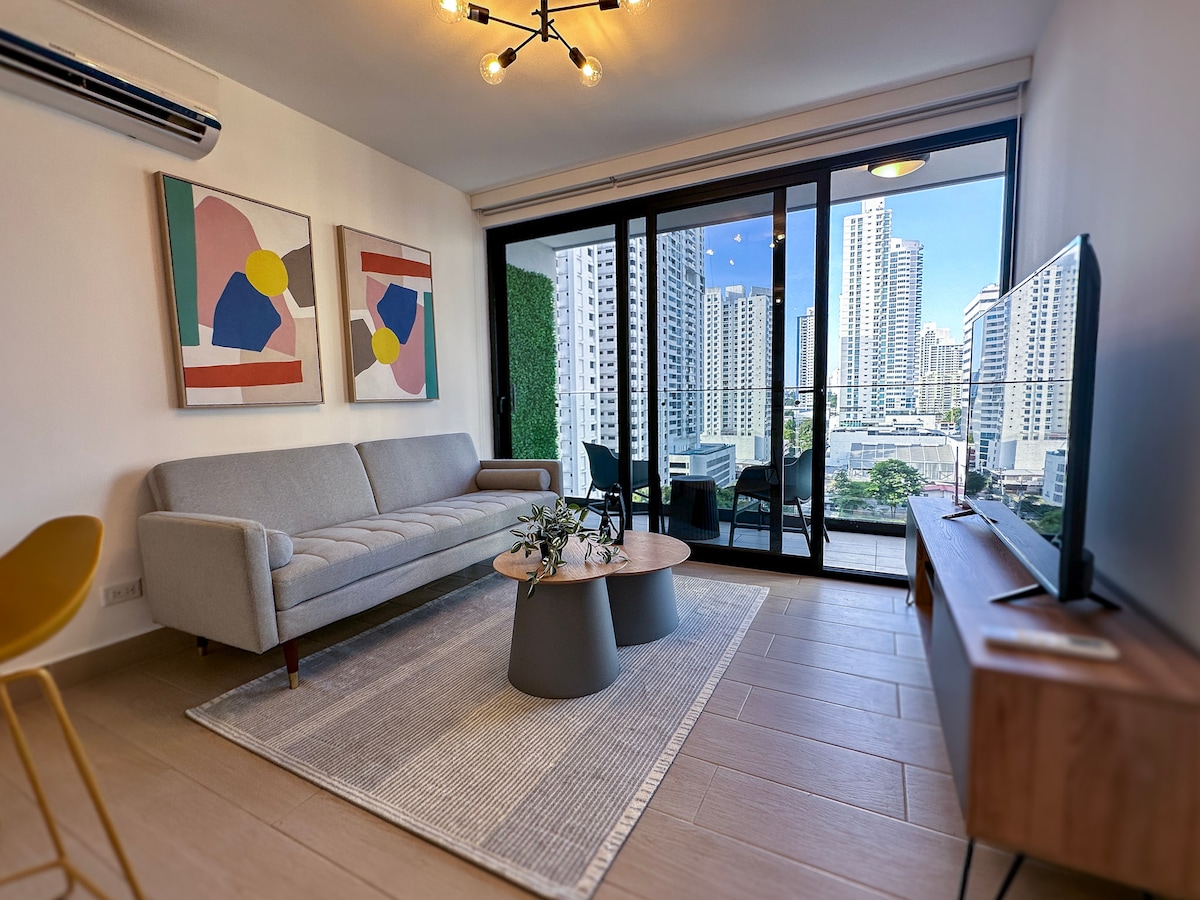 A modern living area is showcased, featuring a light gray sofa positioned across from a flat-screen TV on a wooden console. Two abstract art pieces adorn the walls. Floor-to-ceiling windows offer views of the city skyline, enhancing natural light in the space.