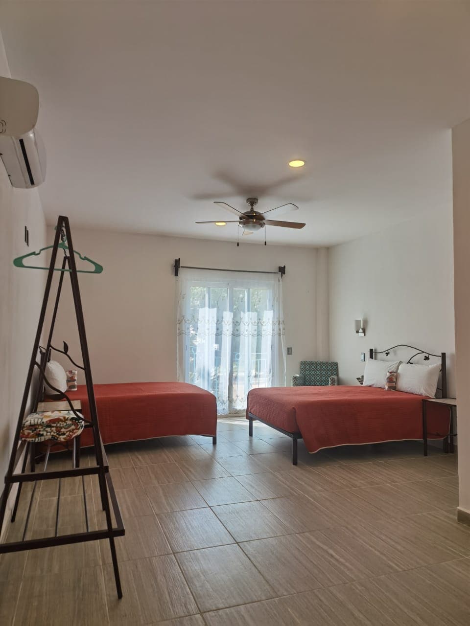 A bright room is furnished with two spacious beds dressed in rust-colored covers. A window with sheer curtains allows natural light to fill the space. A ceiling fan is visible above, and a clothing rack is positioned to one side, enhancing the room's functionality.