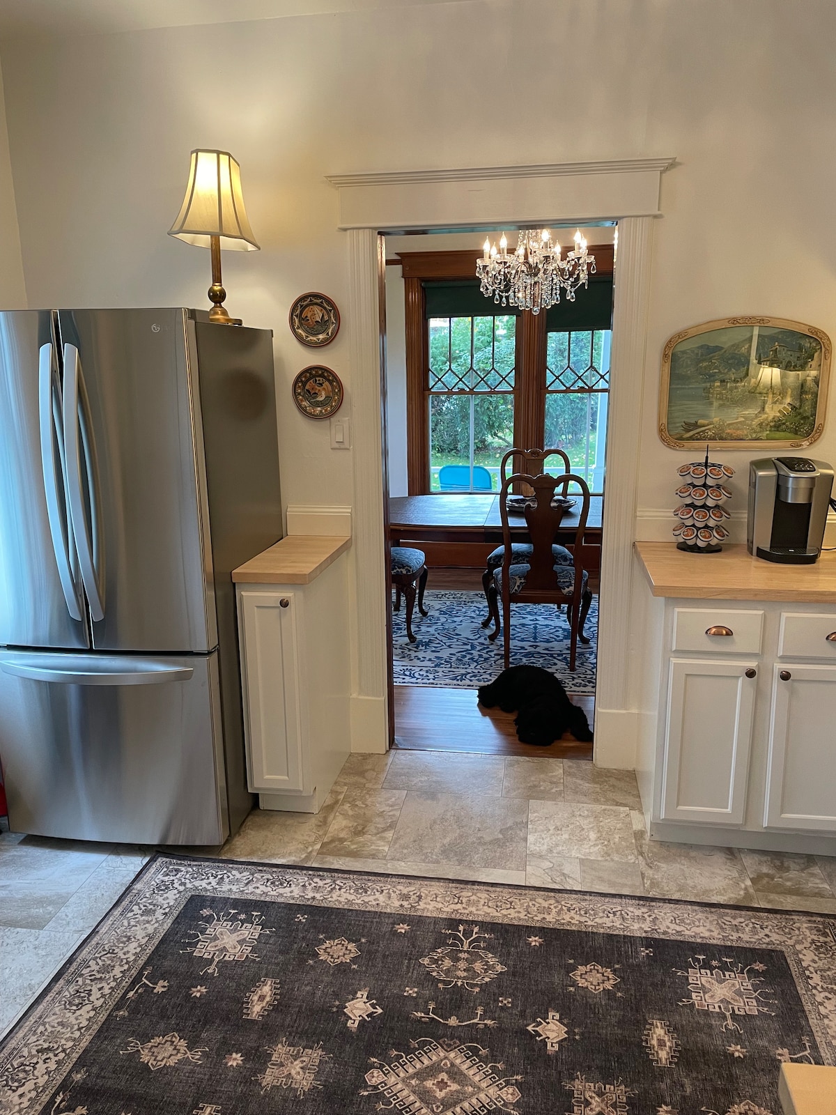The kitchen features modern stainless steel appliances, including a large refrigerator and a coffee machine. Natural light filters through a doorway leading to a dining area furnished with a wooden table and chairs. A decorative chandelier adds an elegant touch to the space.