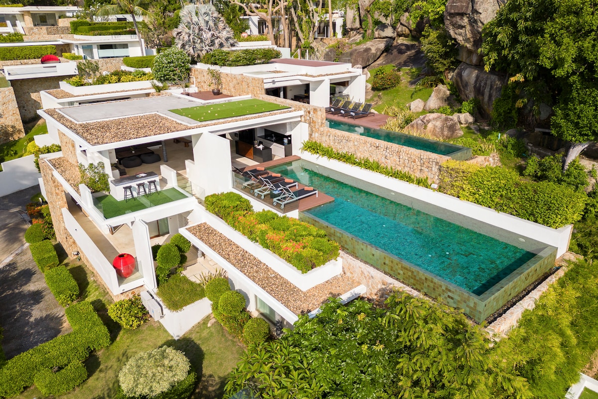 An aerial view captures the modern villa, highlighting a lush landscape with manicured gardens and rocky outcrops. The property features an inviting infinity pool beside a spacious terrace, equipped with sun loungers and shaded seating, seamlessly integrating with the surrounding greenery.