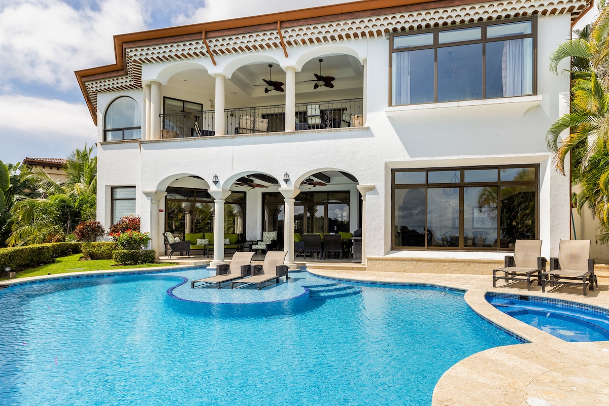 The exterior of the villa is highlighted by an inviting infinity pool surrounded by lounge chairs. Arched columns provide structural elegance, leading to expansive windows that reflect the nearby greenery and sky. Lush landscaping adds to the tranquil outdoor setting.