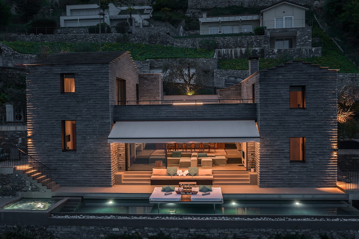 The hillside villa features a modern exterior with wooden cladding, illuminated at dusk. A large terrace showcases a central seating area and walkway extending towards a swimming pool, with views of the landscape and surrounding structures.