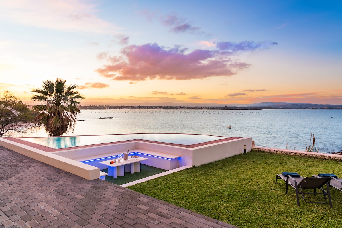 A scenic outdoor space features a curved pool with illuminated edges, a sunken sitting area, and a grassy lawn. Nearby, two lounge chairs provide a space for relaxation, with a view of the tranquil bay and colorful sunset in the background.