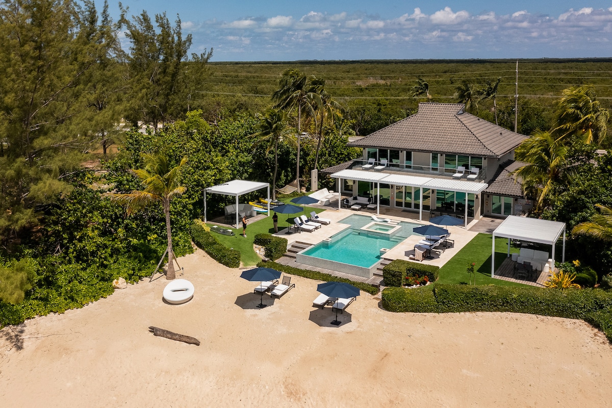 A contemporary villa is set amidst lush greenery, featuring an inviting pool and spacious outdoor seating areas. Sun loungers are positioned near the sandy beach, with views of the ocean beyond. Large windows enhance natural light, creating a seamless connection with the outdoor environment.