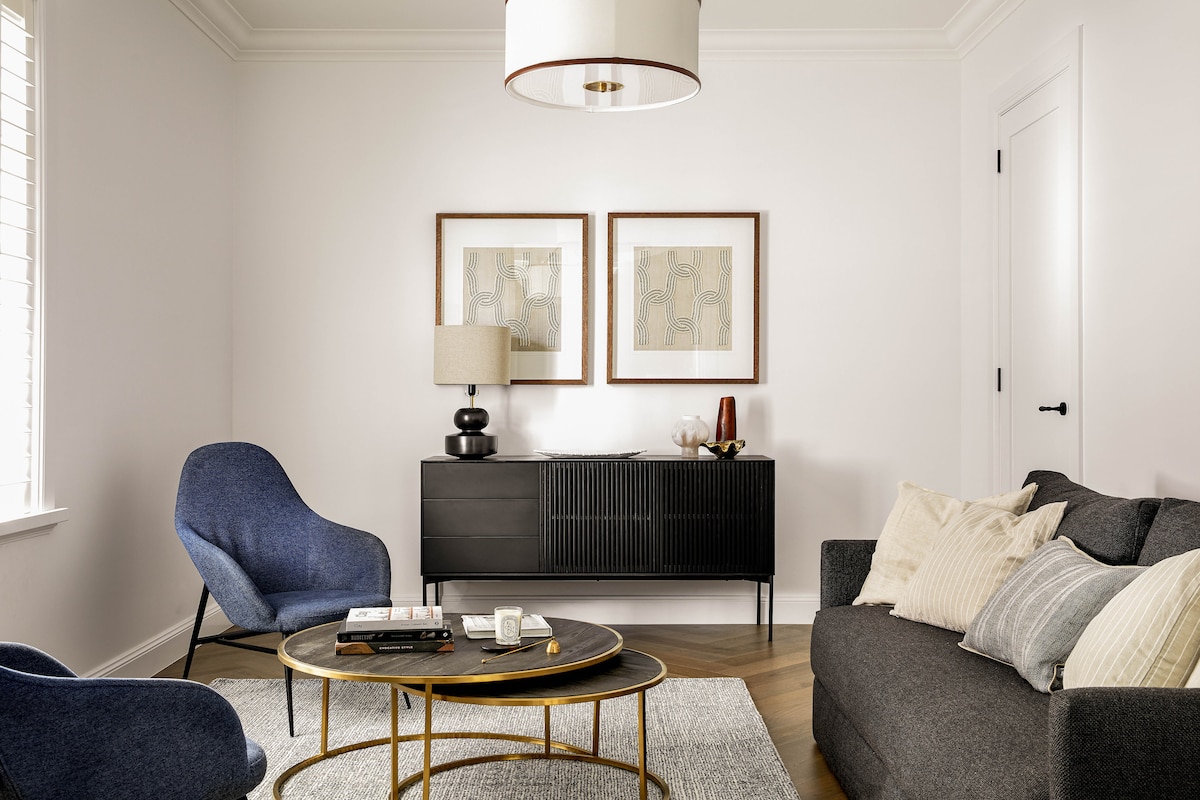 A modern living area features a large gray sofa and blue accent chairs, arranged around a circular coffee table. Artwork adorns the wall above a black sideboard, which holds a lamp and decorative items. Natural light streams in through window blinds.