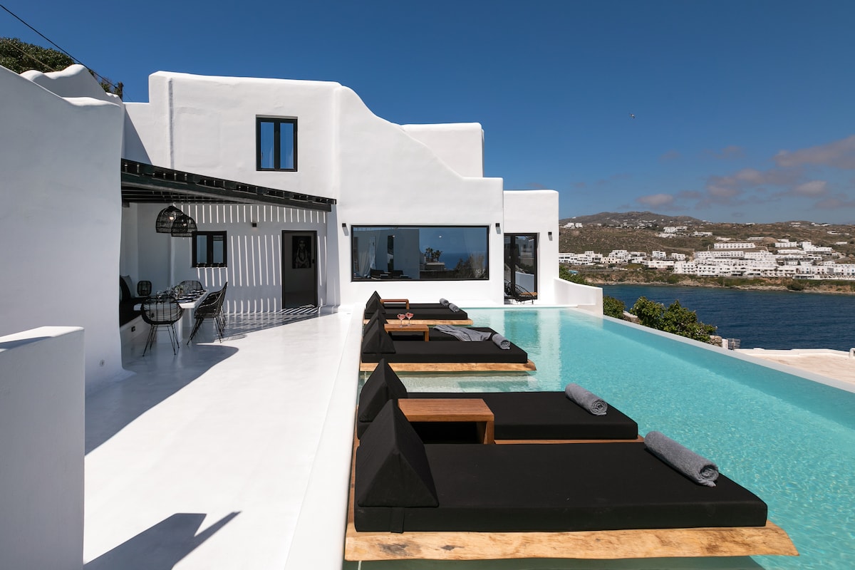 An outdoor terrace features sleek sunbeds positioned near a shimmering pool, which appears to blend into the Aegean Sea. The villa's modern white architecture is complemented by large windows, allowing natural light to illuminate the space. Scenic views of the water and surrounding landscape are visible.