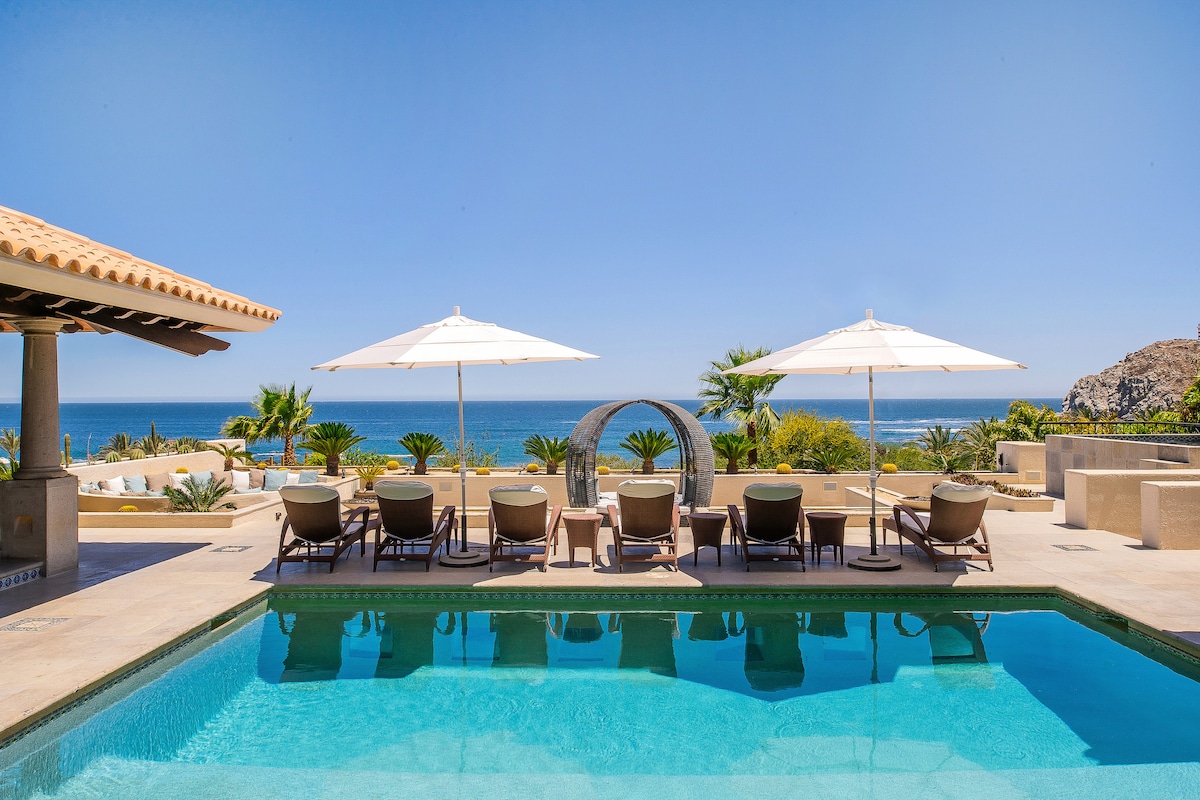 A spacious outdoor area features a crystal-clear swimming pool, surrounded by lounge chairs with large umbrellas for shade. The ocean can be seen in the background, framed by palm trees and landscaped gardens, creating an inviting retreat for relaxation and gathering.