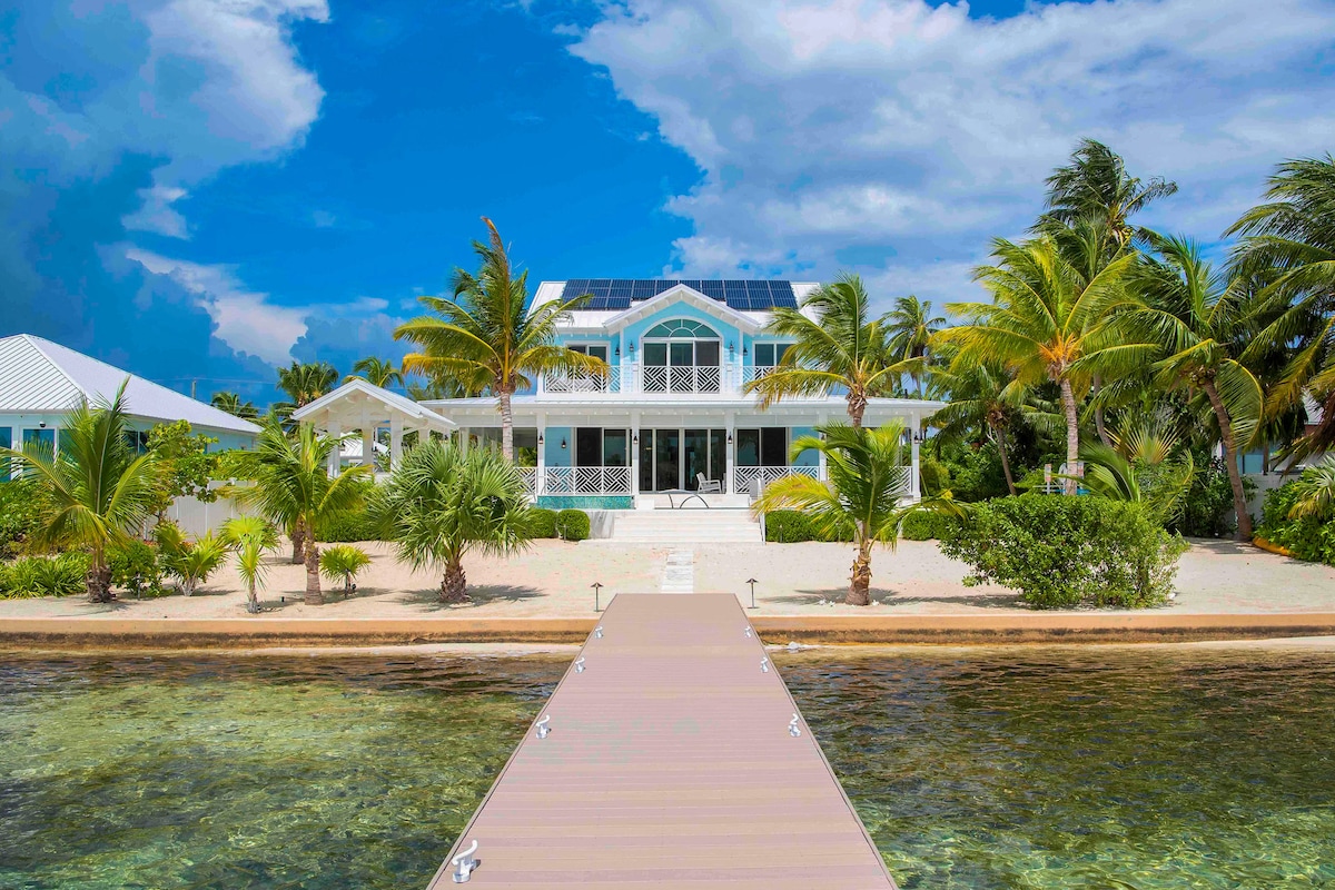 The waterfront beach villa is set against a vibrant sky, with palm trees dotting the landscape. A private dock leads to the beach area, and the structure features large windows and a covered entryway that invites natural light into the space.