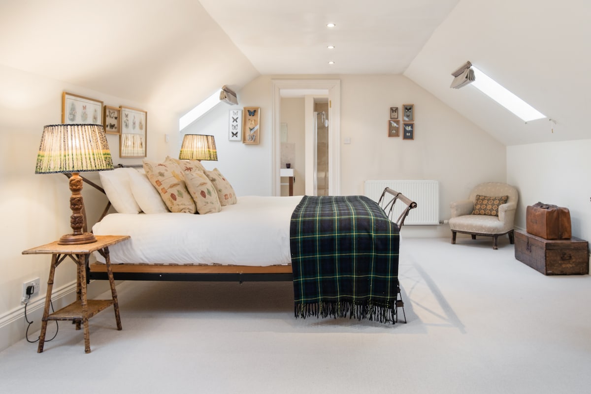 A spacious bedroom features a large bed adorned with pillows and a tartan throw. Two elegant lamps provide soft lighting, while a cozy armchair and vintage suitcase add charm. Skylights allow natural light to fill the room, enhancing its neutral décor.