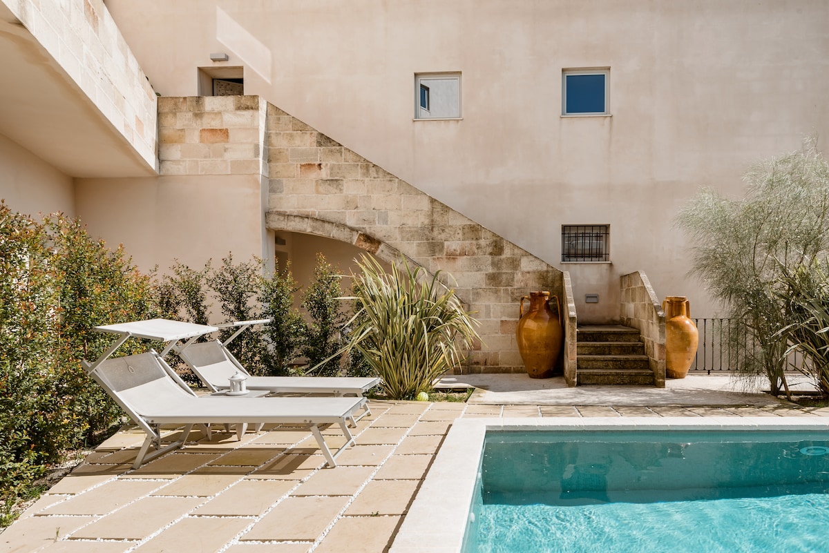A well-maintained outdoor area features a spacious pool surrounded by light-colored stone tiles. Two sun loungers are positioned beside the pool. A set of stairs leads to an upper level, flanked by lush greenery and decorative terracotta pots.