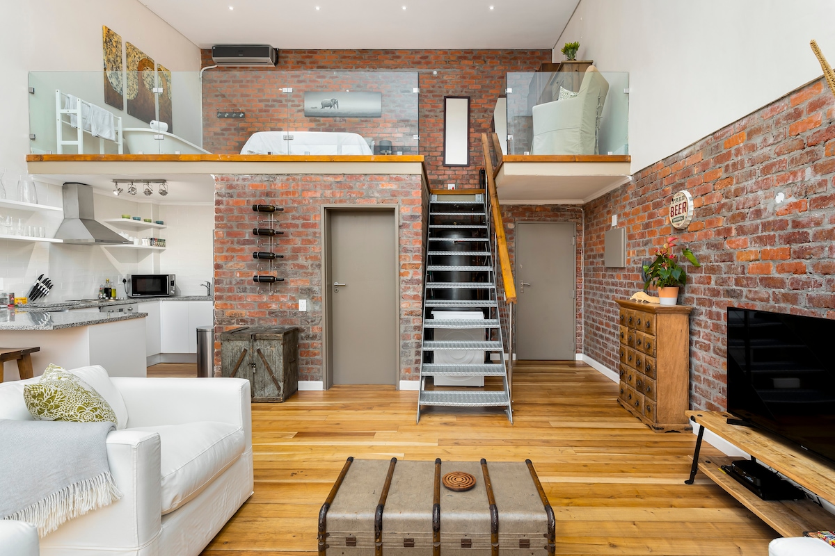 The open-plan living area features exposed brick walls and wooden flooring, creating a warm ambiance. A staircase leads to the mezzanine level, while the space includes a comfortable couch, a flat-screen TV, and a kitchen area equipped with modern appliances.
