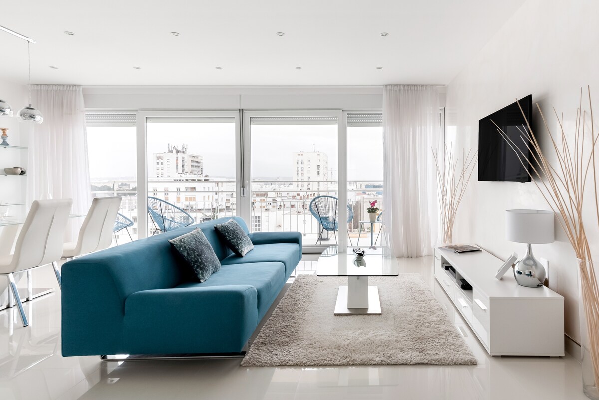 A modern living area features a spacious blue sofa and a glass coffee table, complemented by a light rug. Large windows provide expansive city views, with sheer curtains allowing soft natural light. A television mounted on the wall and minimalistic decor elements enhance the contemporary design.
