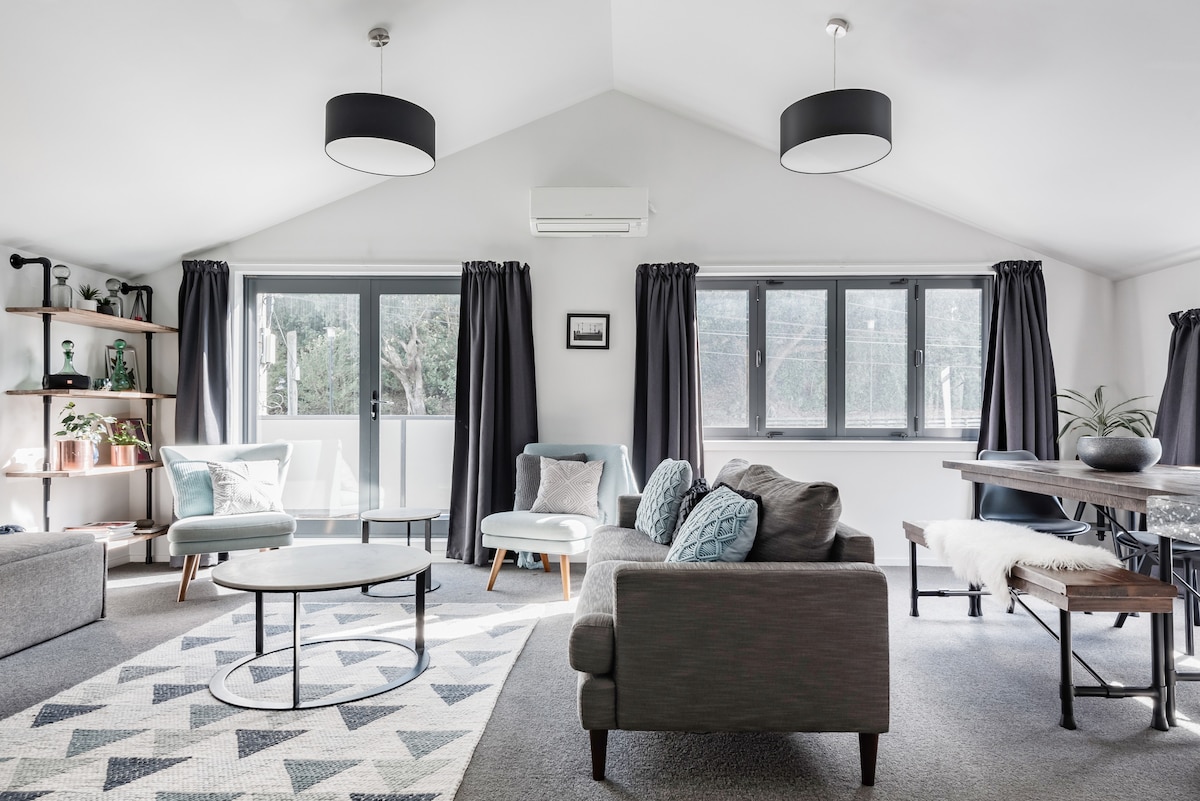 The spacious living area features a combination of modern furnishings, including a comfortable sofa and accent chairs. Large windows allow natural light to fill the room, while dark curtains provide privacy. A round coffee table sits on a patterned area rug, complemented by a dining table nearby.