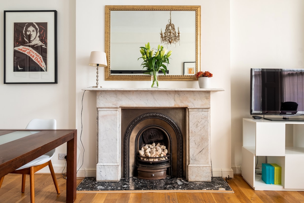 A marble fireplace is framed by a decorative mirror and a ceiling pendant light. Fresh flowers are set on the mantel above the fireplace. A TV is positioned on a white shelf beside the fireplace, with a dining table visible in the foreground.
