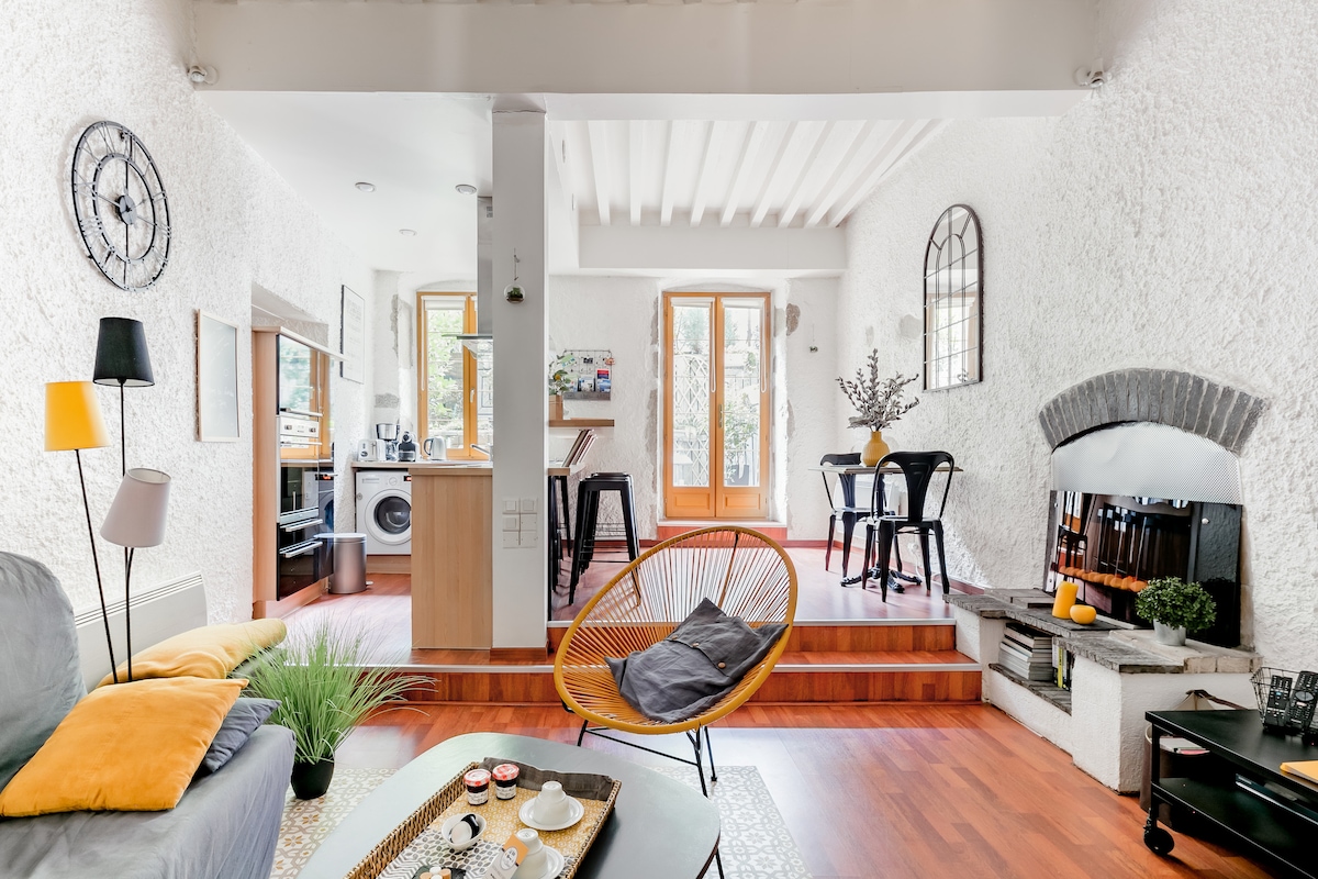 A bright living space features textured white walls and wooden flooring. A comfortable seating area is arranged with a black chair and a colorful accent pillow. A dining area with modern chairs is visible beside large windows, enhancing natural light in the room.