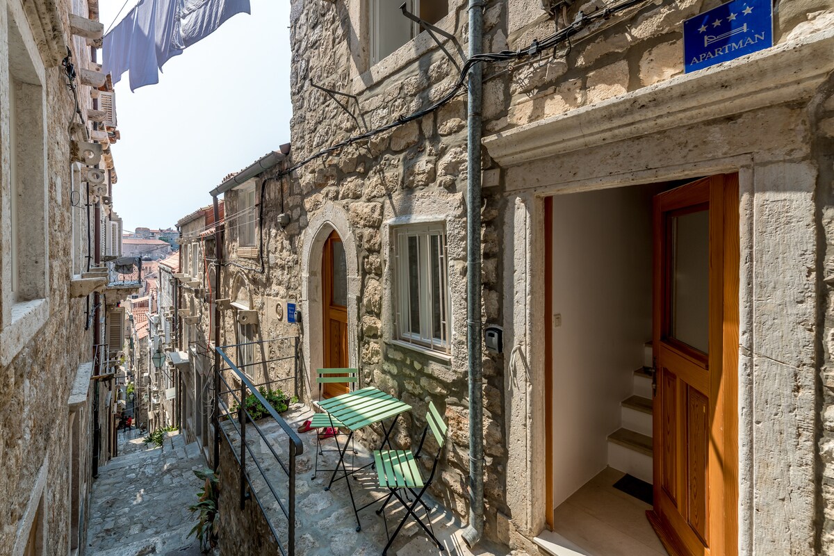A narrow stone street leads to the entrance of a charming apartment, framed by traditional architecture. A small outdoor seating area with green folding chairs and a table is visible, inviting relaxation. Laundry hangs above, adding character to the vibrant atmosphere of the surrounding neighborhood.