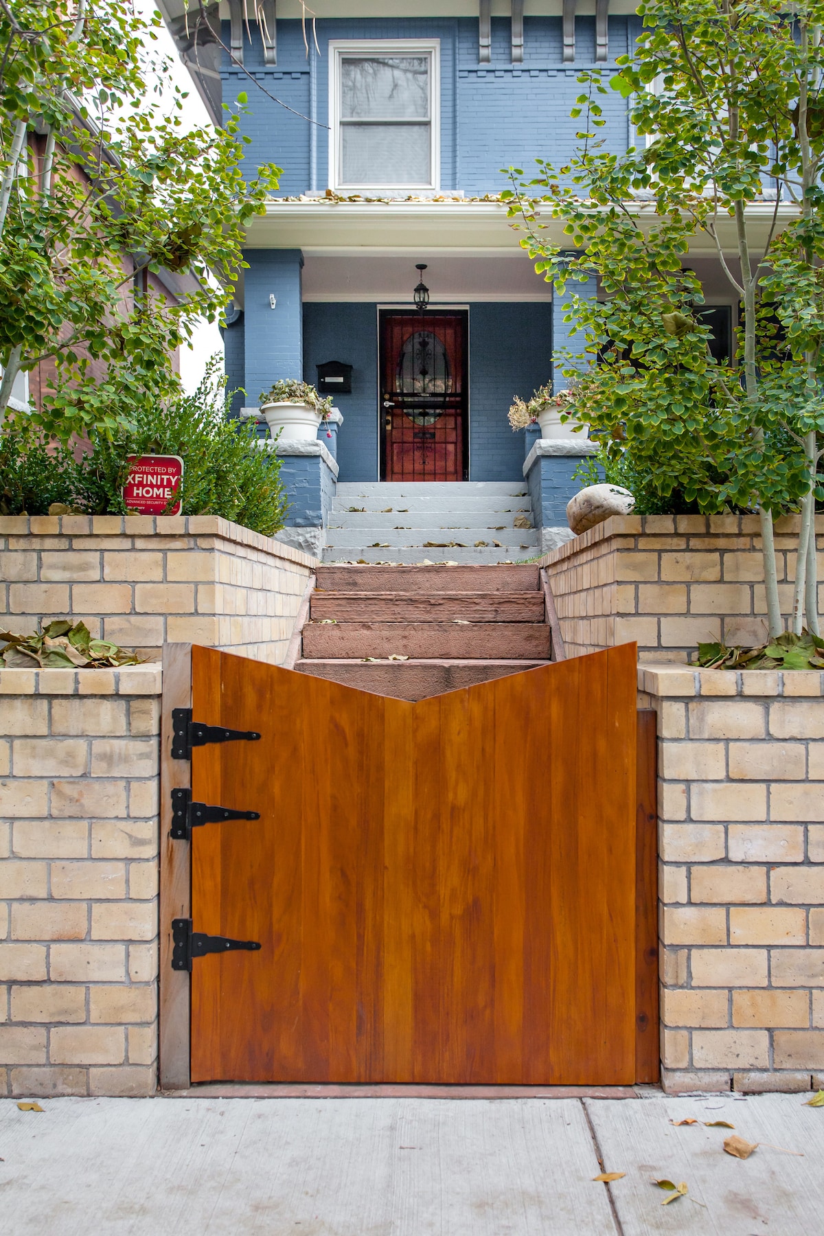 A wooden gate with a distinctive V-shaped top is set within a brick wall, leading to a staircase flanked by plants. The house, with a blue exterior, is visible in the background, showcasing a welcoming entryway.