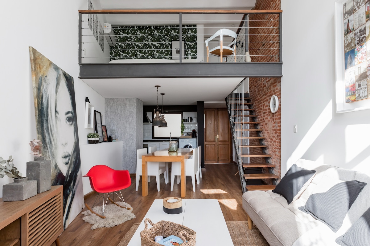 The spacious living area features wood flooring and repointed exposed brick walls. A modern couch and red chair are positioned around a central coffee table. Natural light floods in from large windows, illuminating dining space and the kitchen area, which is accented by a green wall motif.