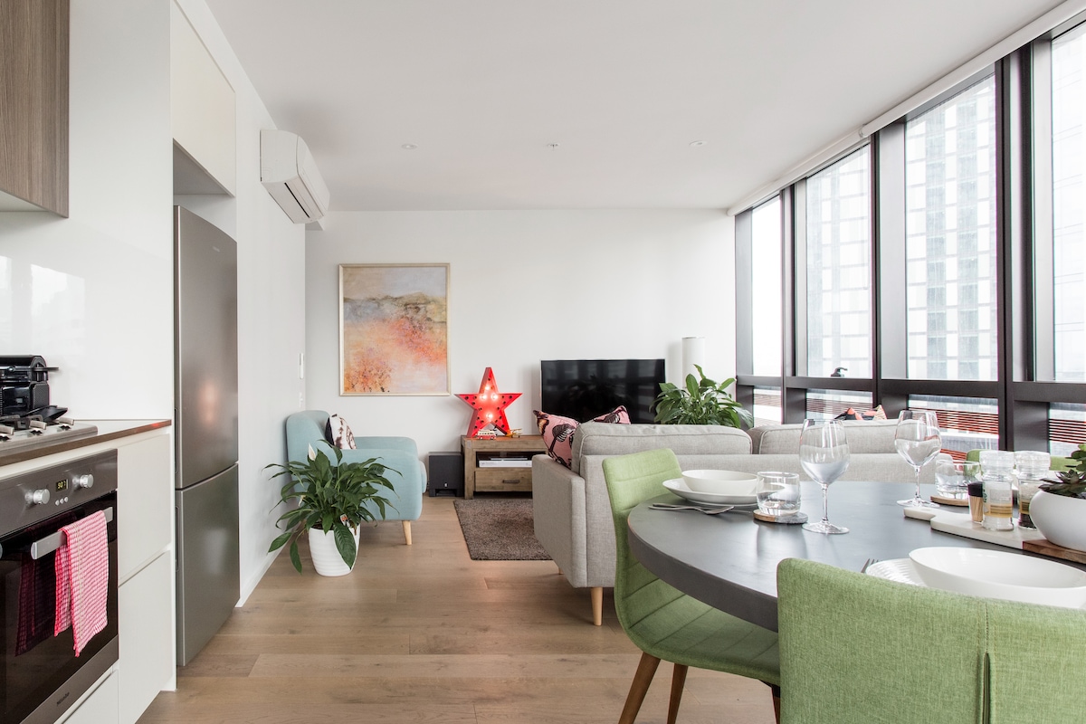 An open-plan living space is showcased, featuring a modern kitchen with stainless steel appliances and a round dining table set for four. A comfortable lounge area includes a large TV and plants, with natural light streaming in through floor-to-ceiling windows.