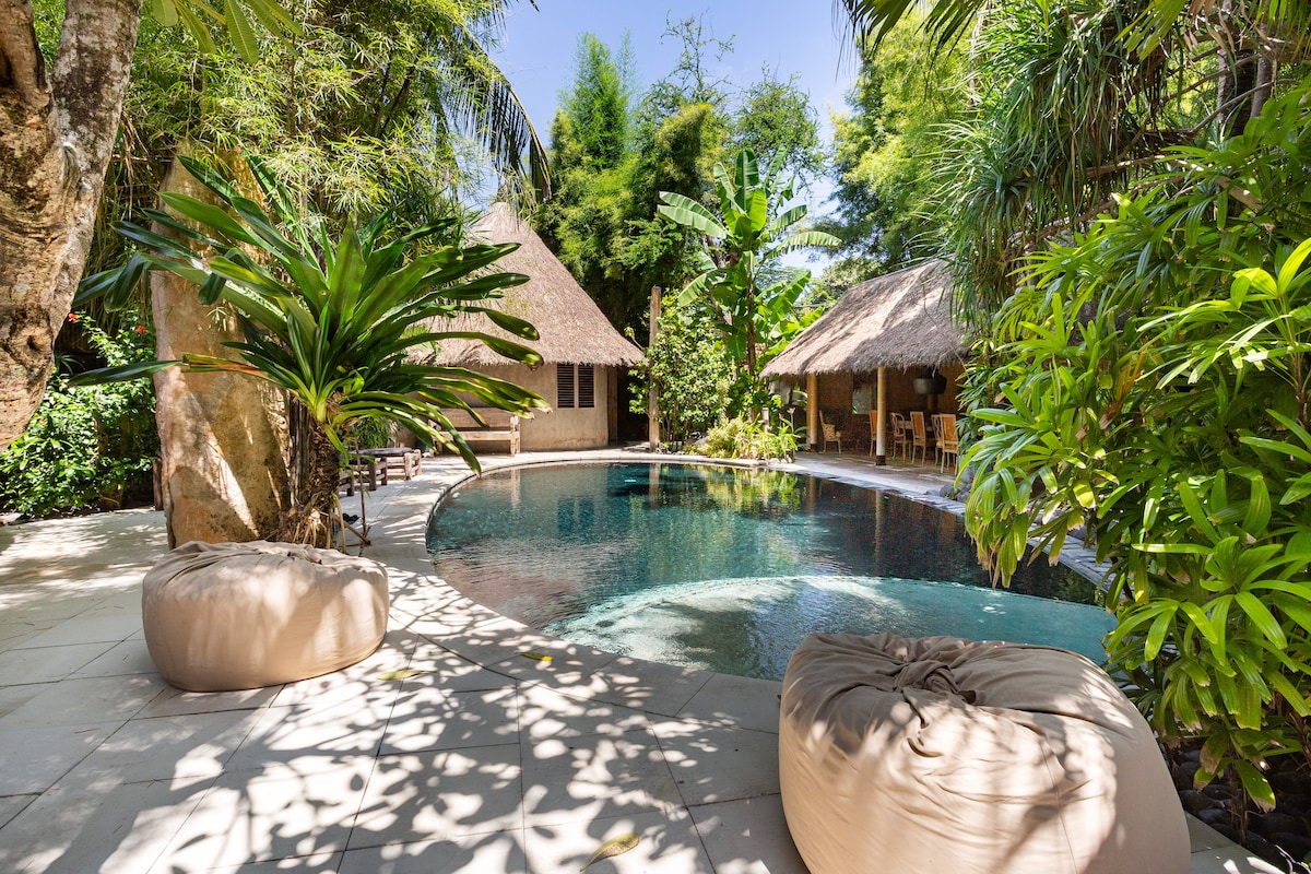 A serene outdoor area features a unique dark pool surrounded by lush tropical plants and trees. Two cozy, oversized bean bags are positioned on the stone paving near the edge of the pool, inviting relaxation in this tranquil, garden-like setting.