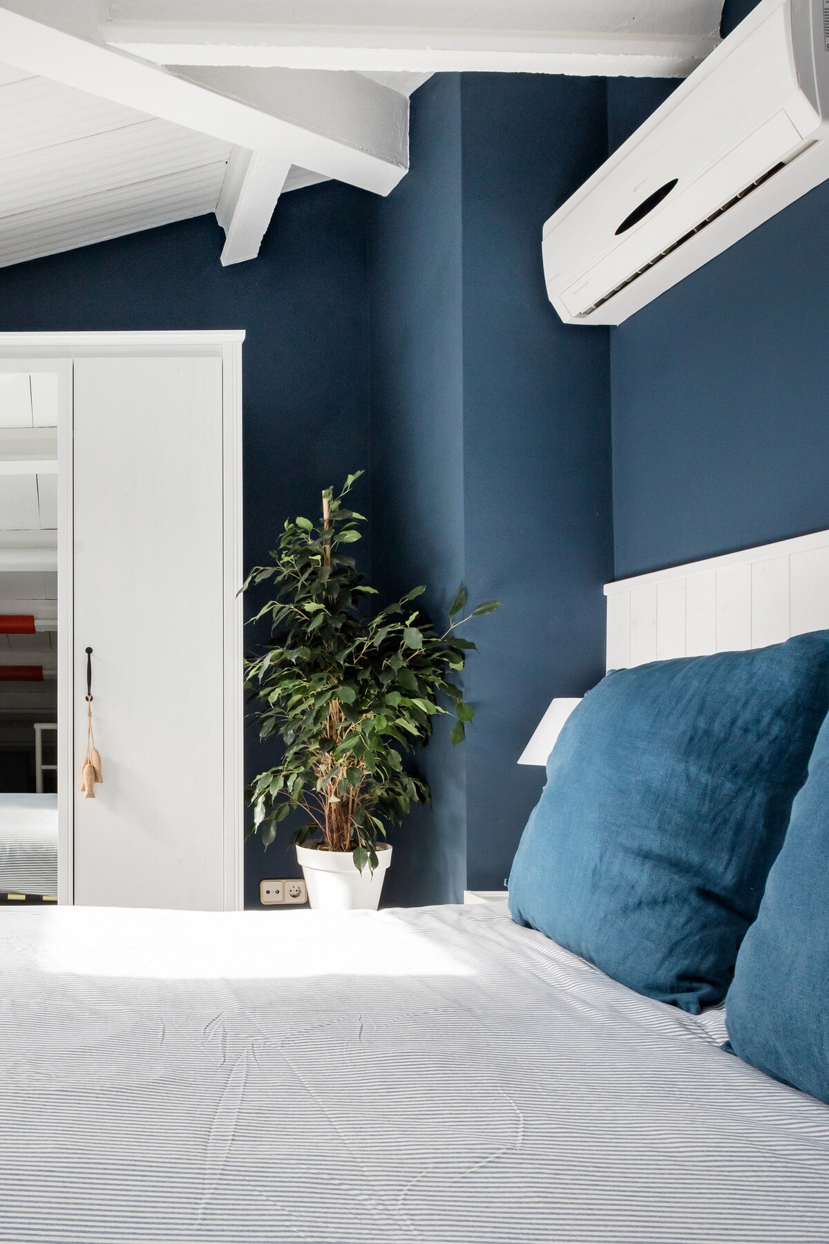 A calming bedroom features a spacious bed with blue cushions against a rich navy wall. A standing plant adds a touch of greenery, while a white wardrobe is subtly positioned nearby. Natural light filters in, enhancing the room's serene ambiance.