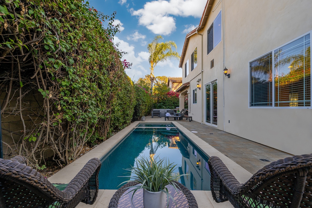 A serene backyard is presented, featuring a narrow pool surrounded by well-maintained greenery. Two woven lounge chairs are positioned nearby, offering a view of the pool and patio area, illuminated by natural light filtering through the leaves of the garden.