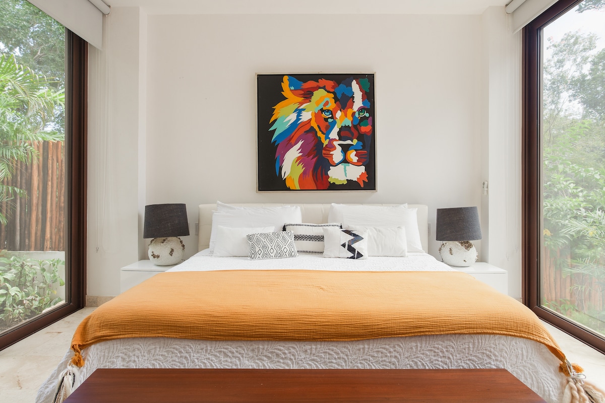 A spacious bedroom features a large bed adorned with a white textured blanket and a vibrant orange throw. Colorful lion artwork enhances the room, complemented by two bedside lamps. Large windows allow natural light and views of greenery to fill the space.