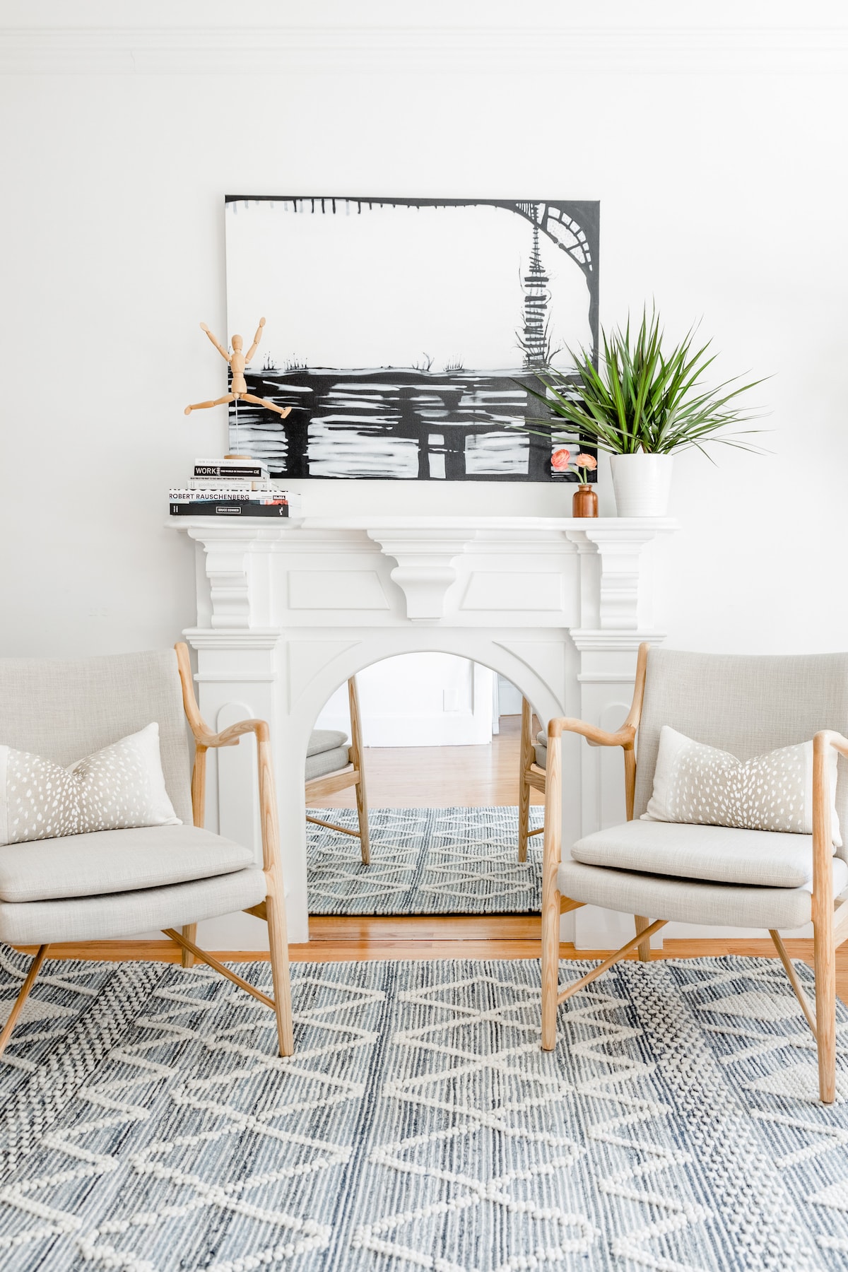 Two light-colored armchairs sit positioned beside a white mantel, which is decorated with a small potted plant and books. Above the mantel, a black-and-white artwork showcases a cityscape. A textured area rug covers the wooden floor, adding warmth to the space.
