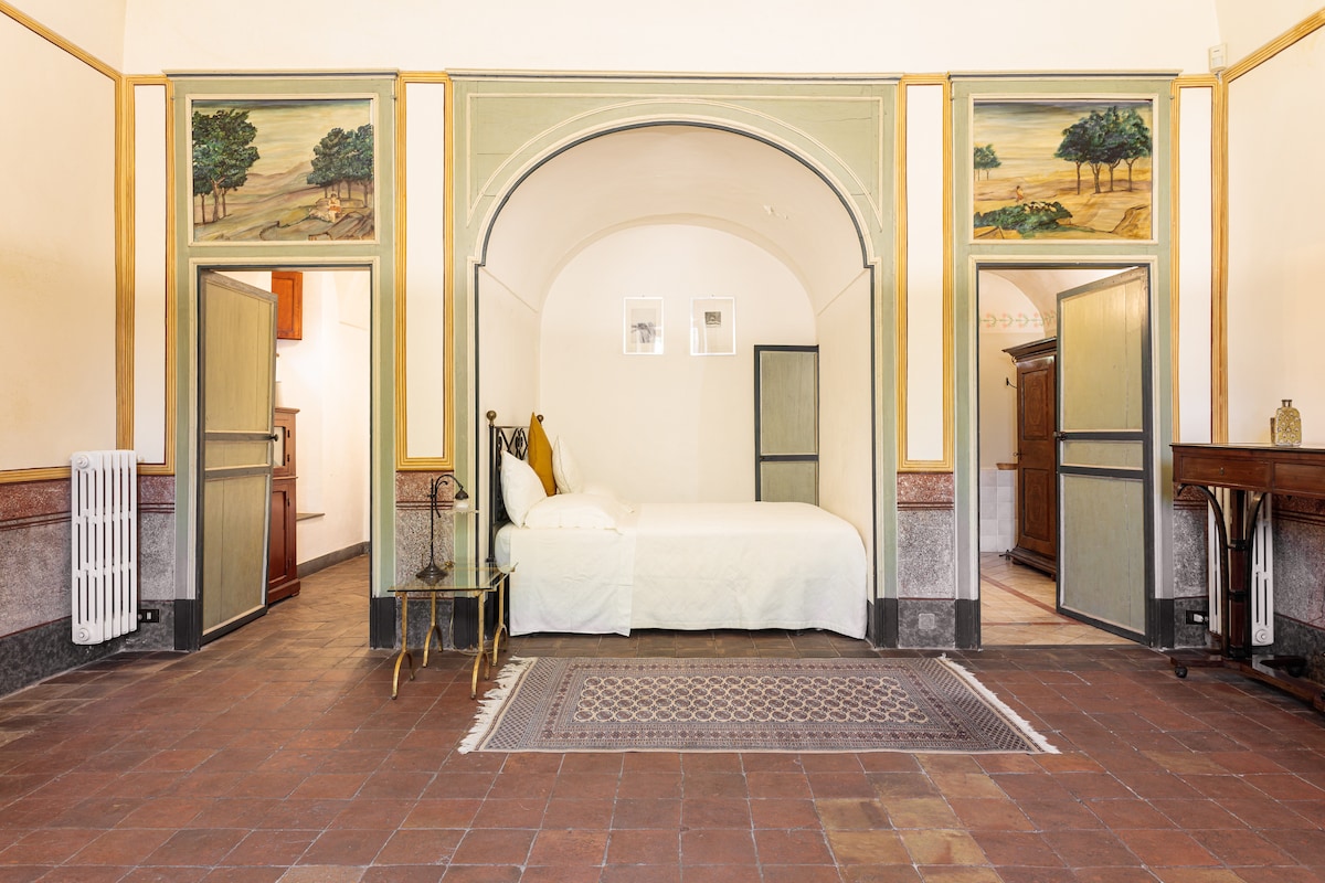 The spacious room features a large alcove with a double bed, framed by intricately painted walls. Light enters through multiple doorways, highlighting a decorative rug on the terracotta floor. A wooden wardrobe and a small side table complete the room's elegant design.