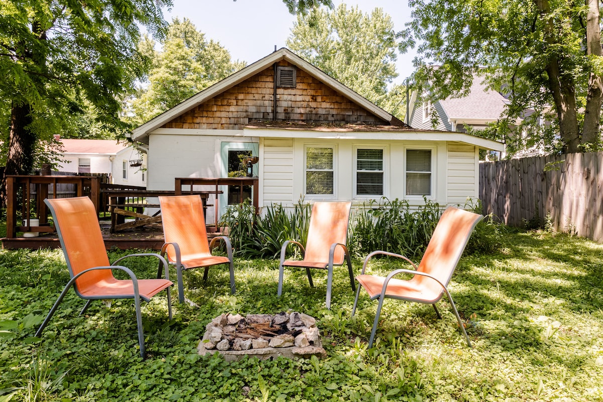 A cozy outdoor area features four orange chairs arranged around a stone fire pit, surrounded by lush green grass and plants. The back of the cottage is seen, with large windows allowing natural light to fill the interior.