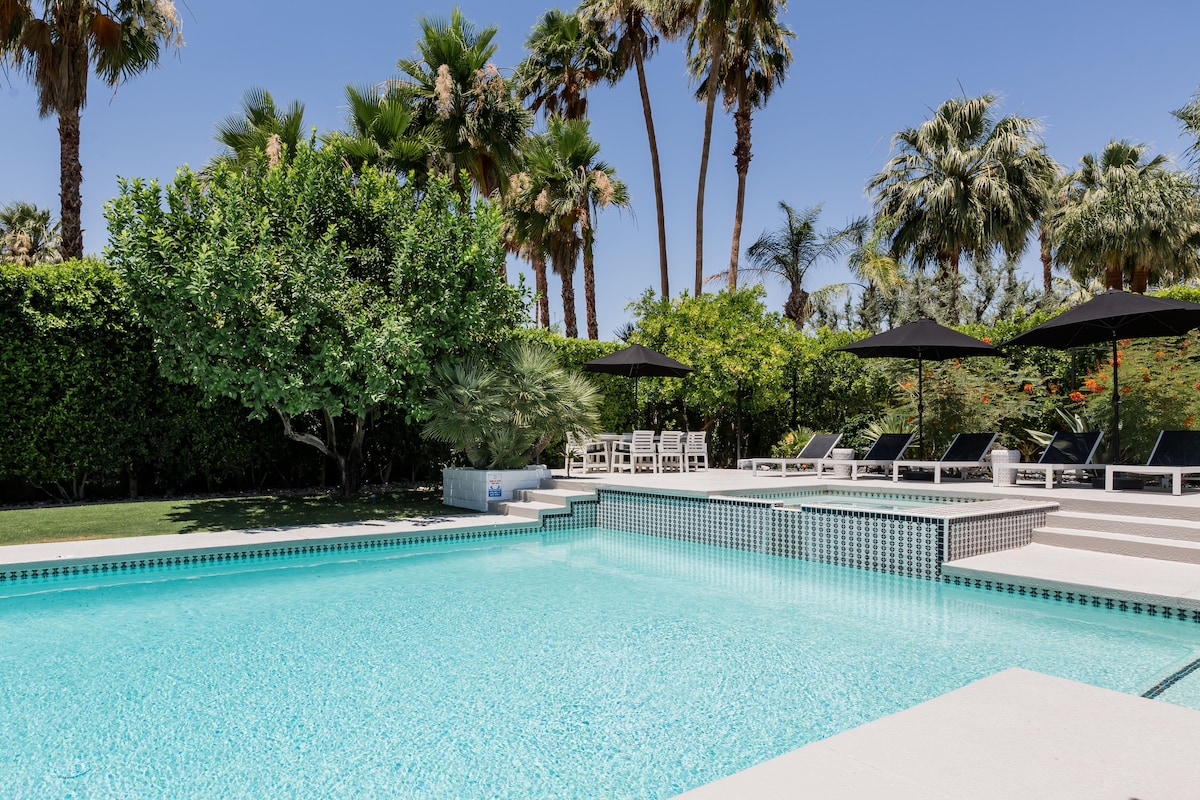 The expansive saltwater pool is surrounded by palm trees and lush greenery. Dark umbrellas provide shade over lounge chairs positioned on the pool deck. A small garden area features citrus trees, enhancing the relaxing atmosphere in the backyard.