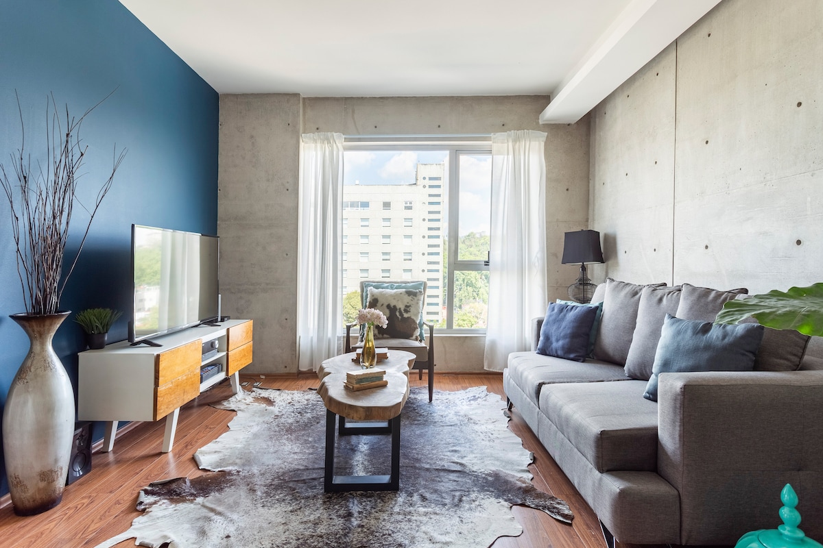 A modern living area is featured, showcasing a light grey sectional sofa positioned around a wooden coffee table. A flat-screen television sits on a sleek cabinet, while large windows with sheer curtains allow natural light to illuminate the space.