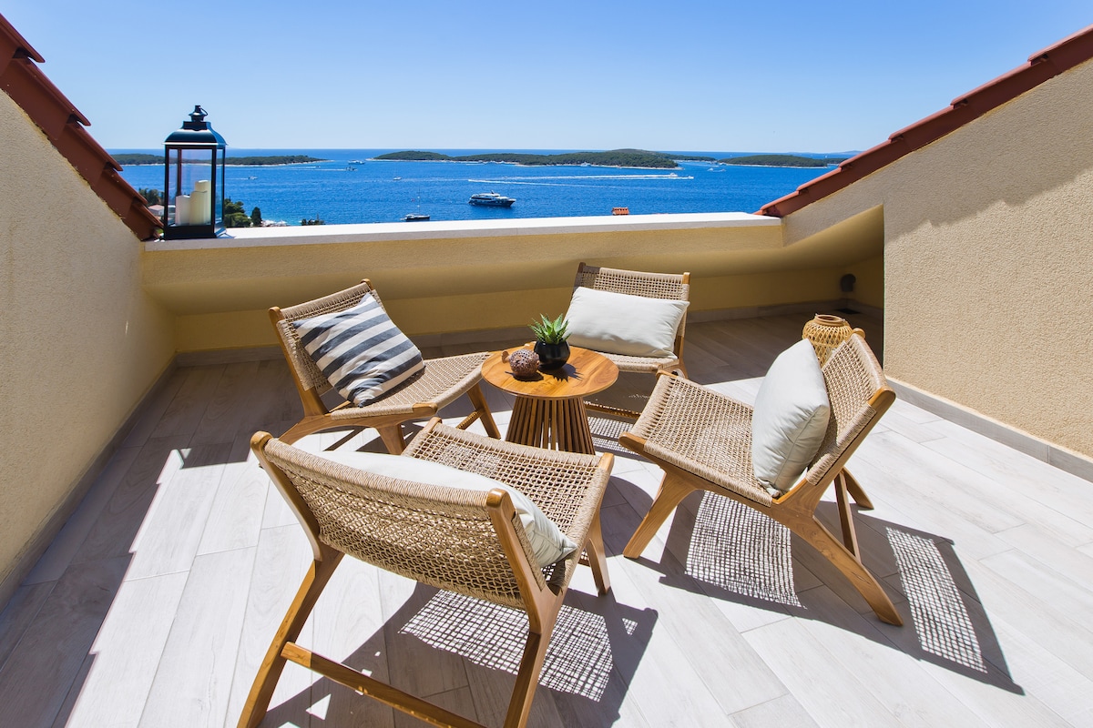 A balcony setting offers a stunning view of the sea, featuring four woven chairs arranged around a circular table. A small plant adorns the table, while the sky above is clear and bright, enhancing the outdoor space's inviting ambiance.