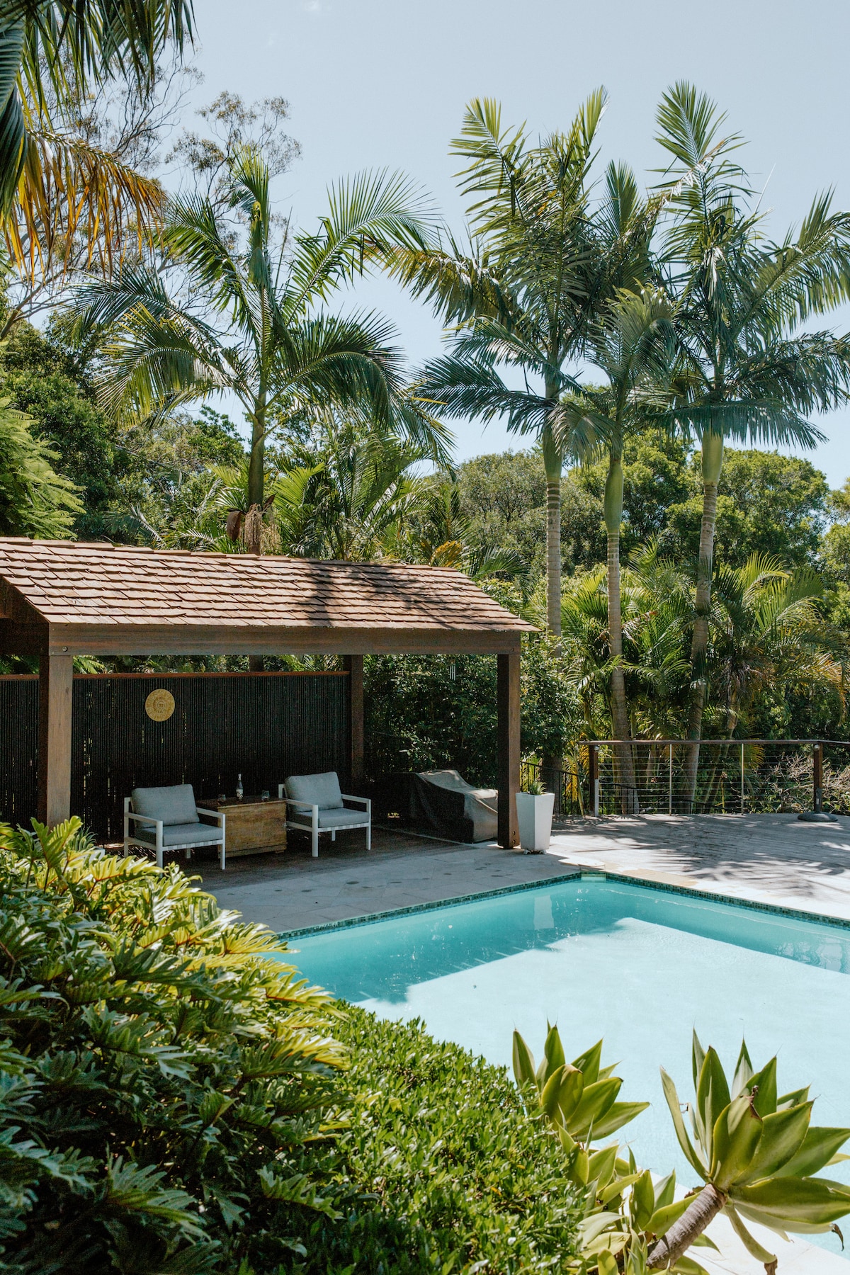 A magnesium pool is framed by lush tropical plants and palm trees, offering a serene atmosphere. The adjacent cabana features two seating areas, inviting relaxation. The clear water reflects the blue sky, enhancing the tranquil setting surrounded by vibrant greenery.