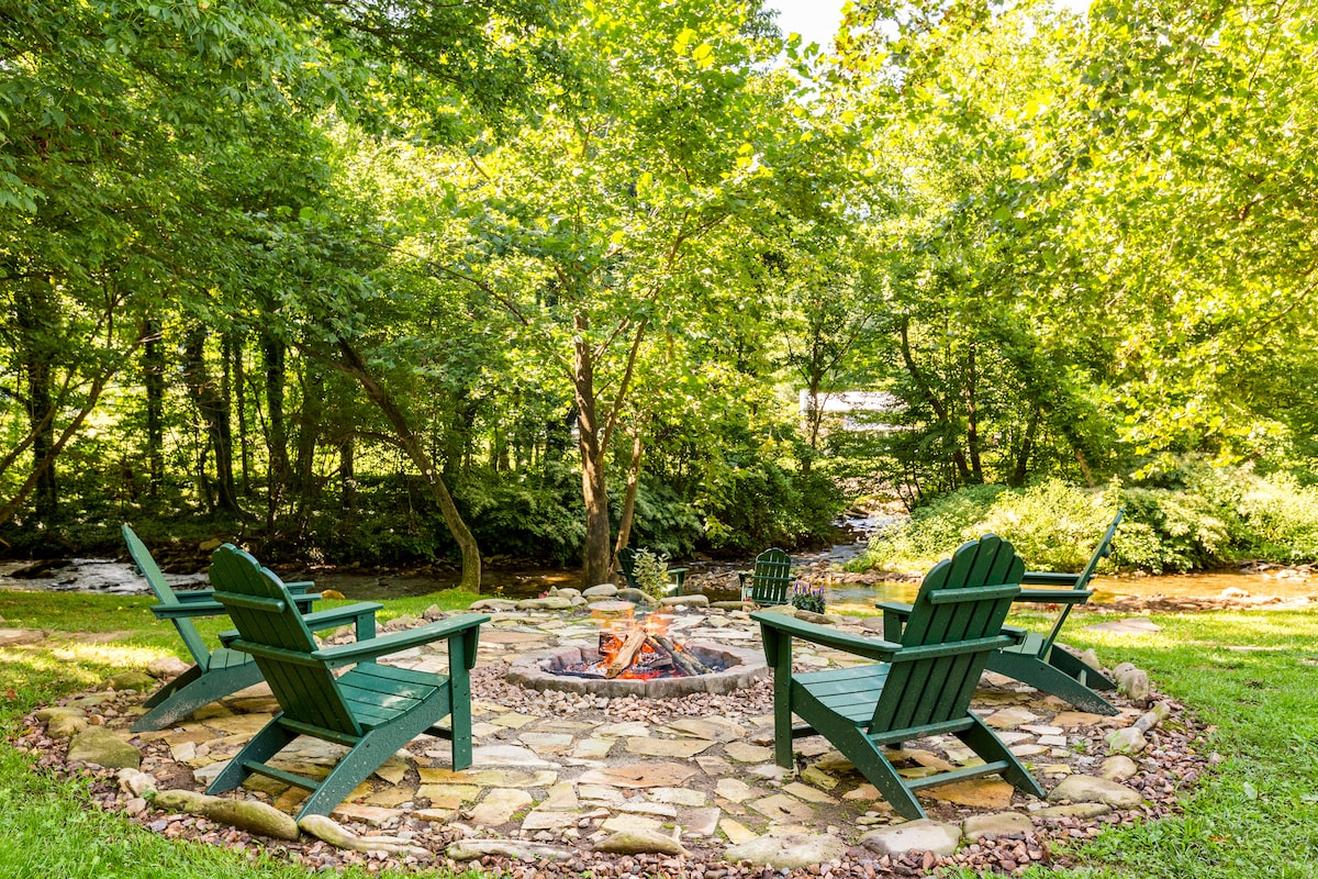 A stone fire pit is surrounded by green Adirondack chairs, inviting relaxation in a natural setting. Lush greenery envelops the area, with a gentle creek flowing nearby, enhancing the serene outdoor space.