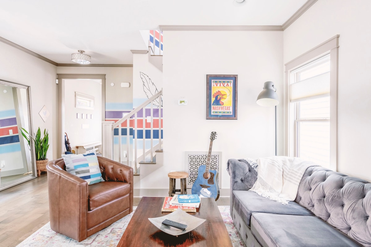 The inviting living area features a cozy sofa adorned with light-colored throw pillows and a leather armchair, complemented by a wooden coffee table. A colorful art piece hangs above the guitar resting beside a small wooden stool, while a vibrant area rug adds warmth to the hardwood floor.