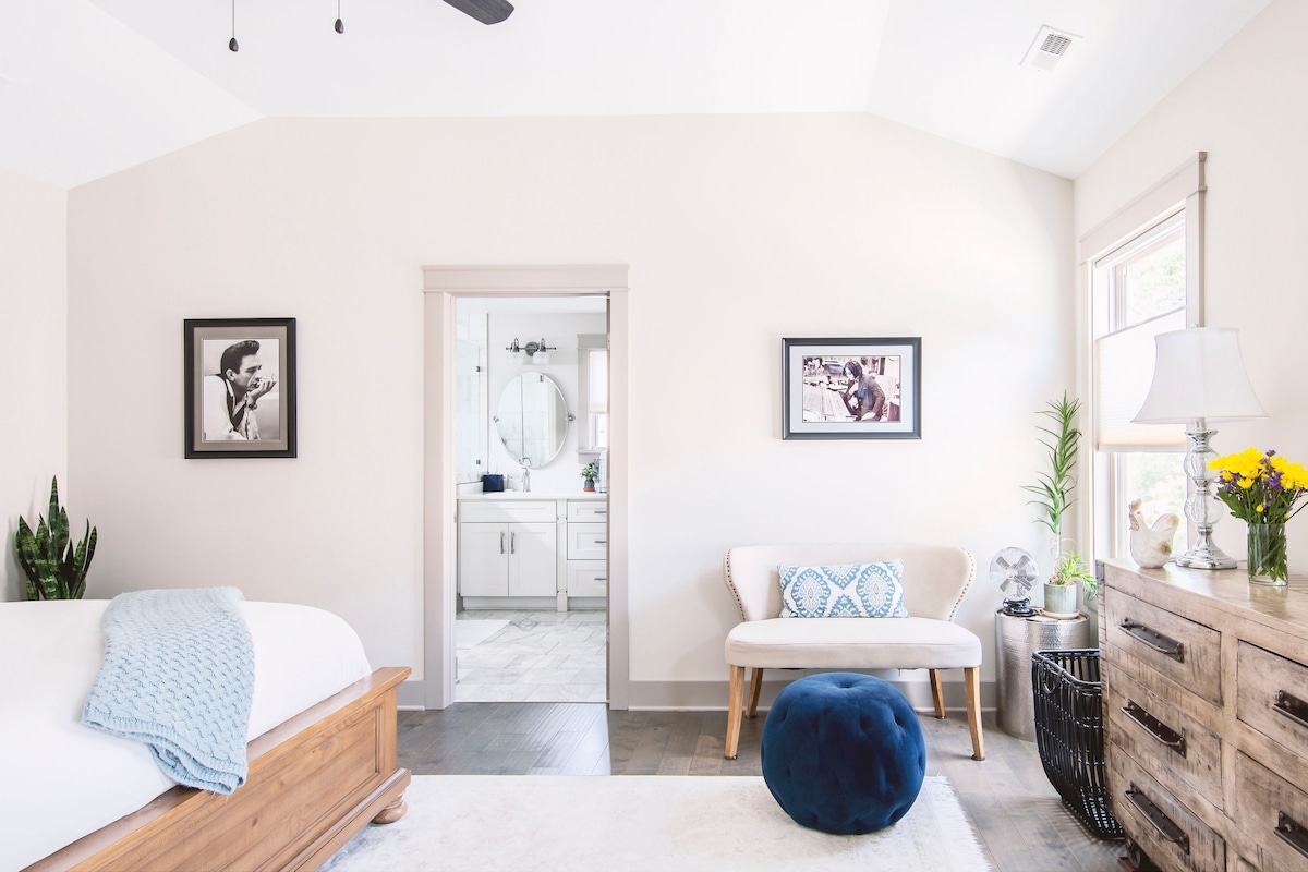 A bright bedroom is showcased, featuring a spacious bed with white linens and a natural wood frame. Nearby, a stylish bench and a blue pouf add seating options. A doorway leads to a modern bathroom, while decorative plants and artwork enhance the overall ambiance.