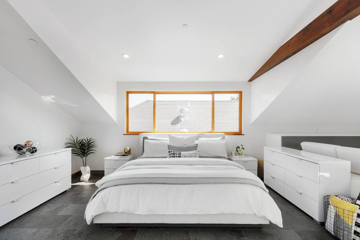 A spacious bedroom is featured, furnished with a large bed dressed in white linens, flanked by white dressers. Natural light fills the room through a wide window, illuminating the sleek surfaces and the minimalist design elements, creating a serene atmosphere.