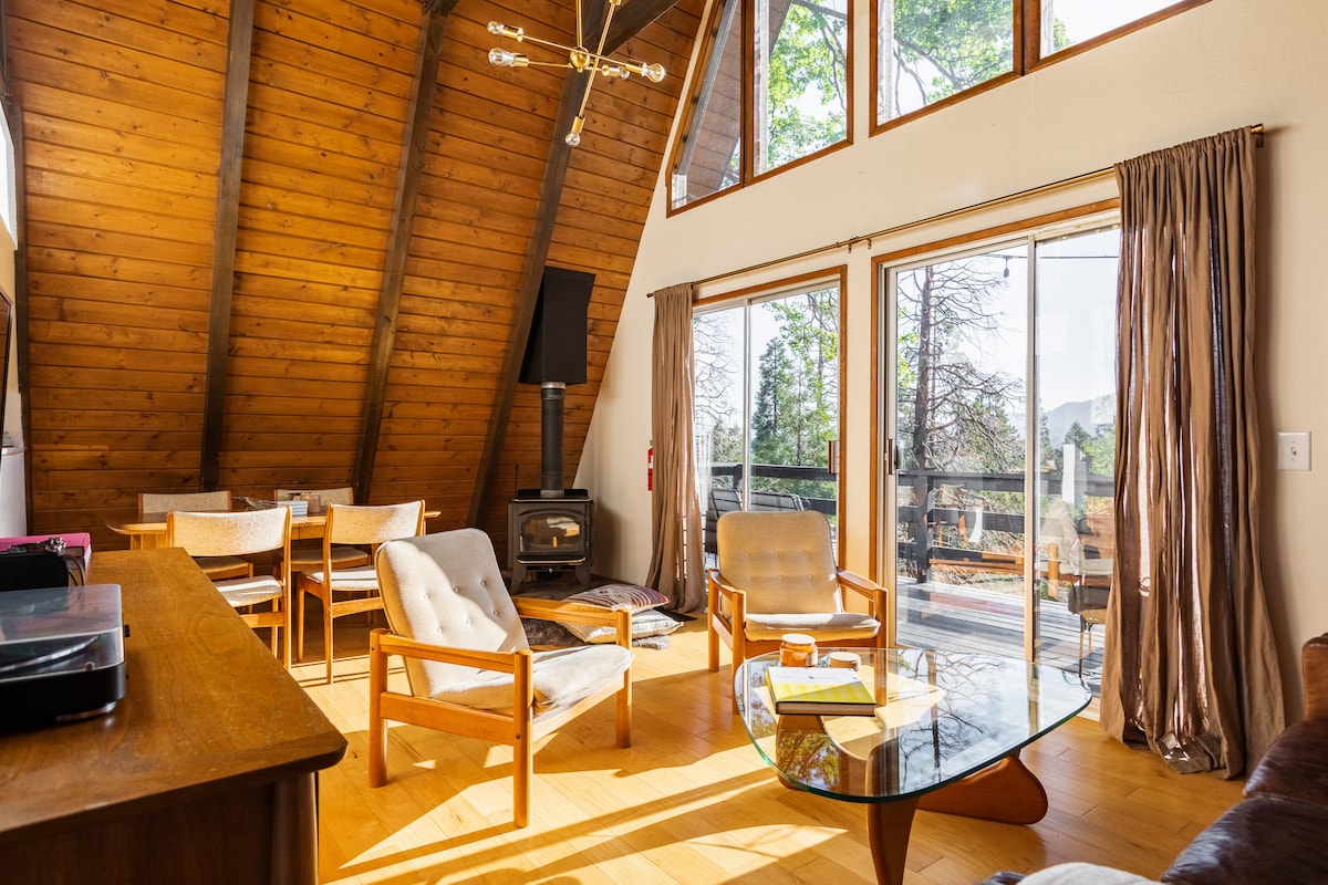 An inviting living area is highlighted by a tall A-frame ceiling and warm wooden walls. Comfortable seating is arranged around a glass coffee table. A large sliding door opens to an outdoor deck, allowing natural light to fill the space.