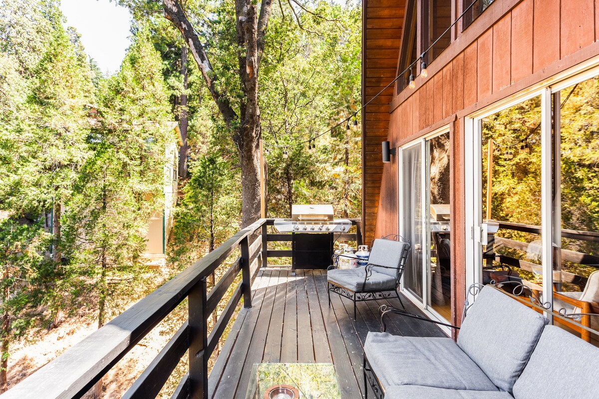 An outdoor deck features comfortable seating arranged around a glass-top table. Lush greenery surrounds the area, providing a sense of privacy. A barbecue grill is visible at the far end of the deck, enhancing the potential for outdoor dining and relaxation in nature.