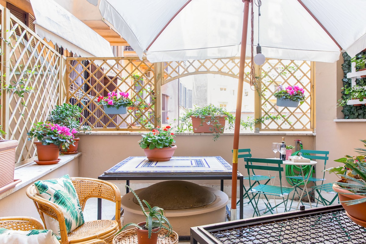 A peaceful patio area is highlighted by a large umbrella providing shade over a dining table with green chairs. Flower pots filled with vibrant blooms are arranged along the trellis, while a cozy seating area features rattan chairs and greenery, creating an inviting outdoor space.