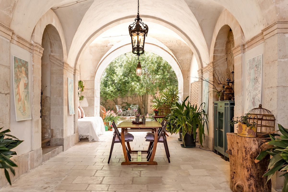 A charming open space features arched stone ceilings with elegant lantern lighting. A wooden table surrounded by chairs is placed at the center, while potted plants enhance the natural ambiance. Beyond, a lush garden is visible, offering a serene view.