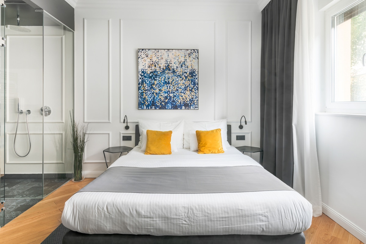 The sleeping area features a spacious bed dressed in white linens with a gray throw. Two yellow accent pillows add a pop of color. A large artwork is displayed above the bed. A glass shower is visible in an adjoining area, with plants positioned nearby.