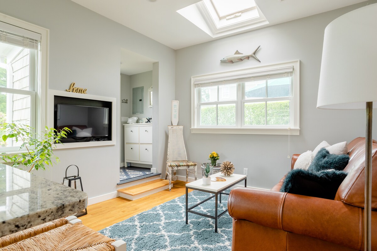 A welcoming living area features a cozy leather sofa paired with a decorative coffee table atop a textured area rug. A skylight and large windows provide natural light, while a flat-screen TV is mounted on the wall. Steps lead to a bright bathroom space.