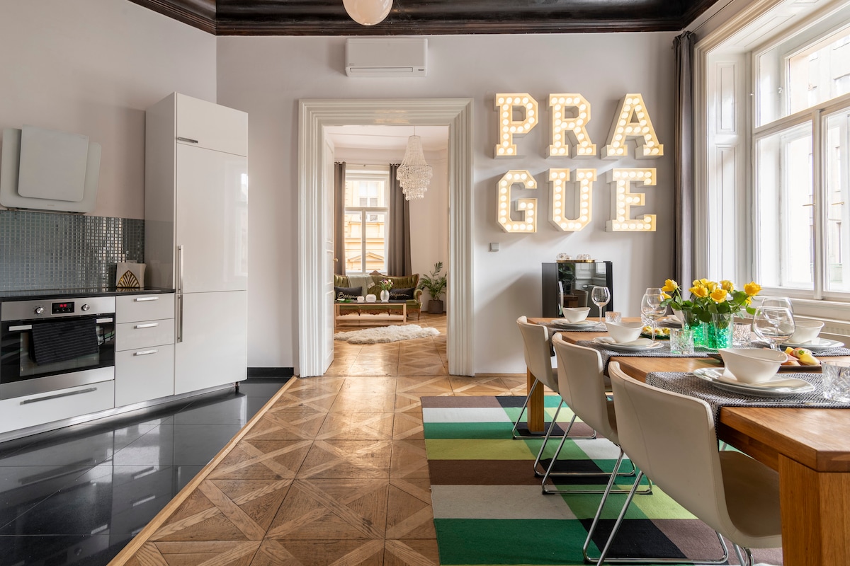 A dining area is featured with a wooden table set for a meal, complemented by modern chairs. In the background, a kitchenette is visible, equipped with sleek appliances. A bright sign spelling 'PRAGUE' adds a playful element to the room, enhancing its welcoming ambiance.