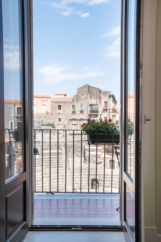The image displays an open balcony door, revealing a view of the Roman Theater and surrounding buildings. Flower boxes adorn the balcony, adding a touch of greenery against the historic backdrop. A clear blue sky is visible, enhancing the inviting feel of the space.