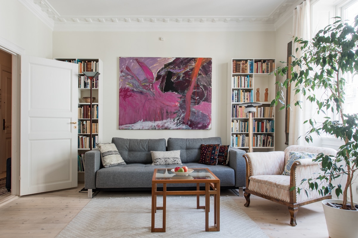 A spacious living room features a large grey sofa adorned with decorative pillows. A vibrant abstract painting is displayed above the couch. A wooden coffee table sits in the center, surrounded by two bookshelves filled with books, while a green potted plant adds a touch of nature.